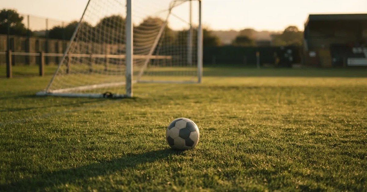 Ein Fußball liegt vor einem leeren Tor auf dem Rasen, das Netz hängt unbewegt, im Hintergrund ein verschwommener Zaun in warmem Abendlicht.