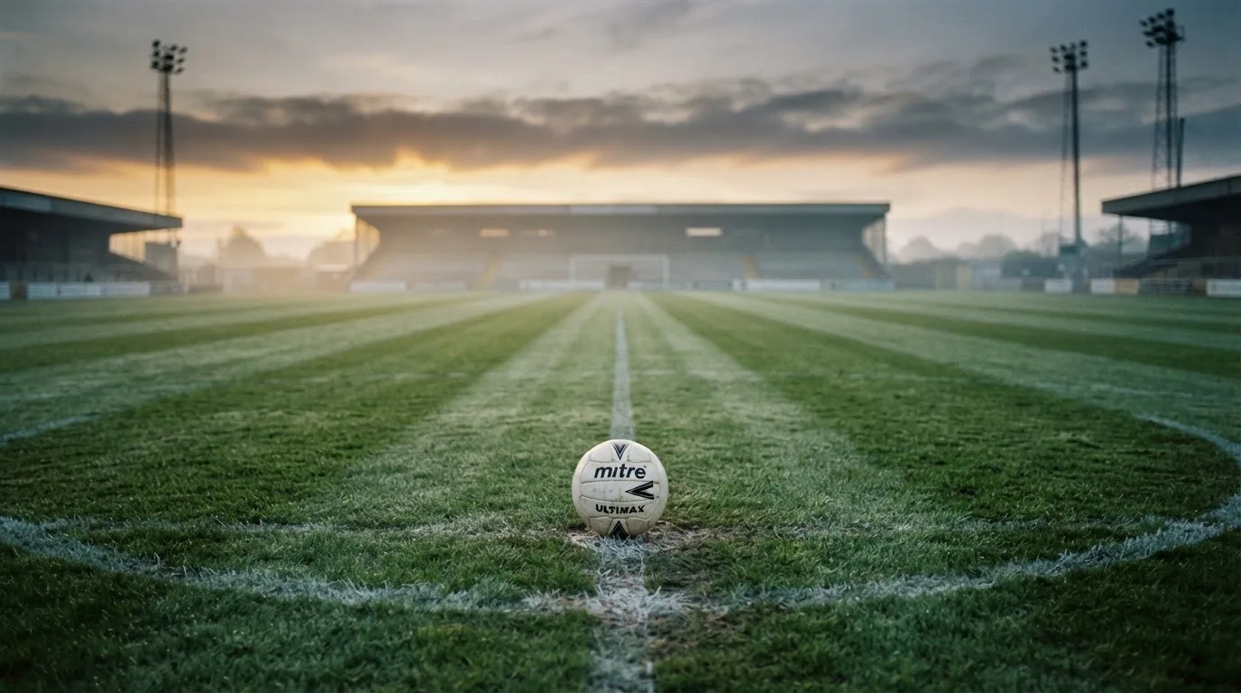 Ein Fußball liegt auf dem Mittelkreis eines leeren Rasenplatzes am frühen Morgen, im Hintergrund eine ferne Tribüne im Dunst.