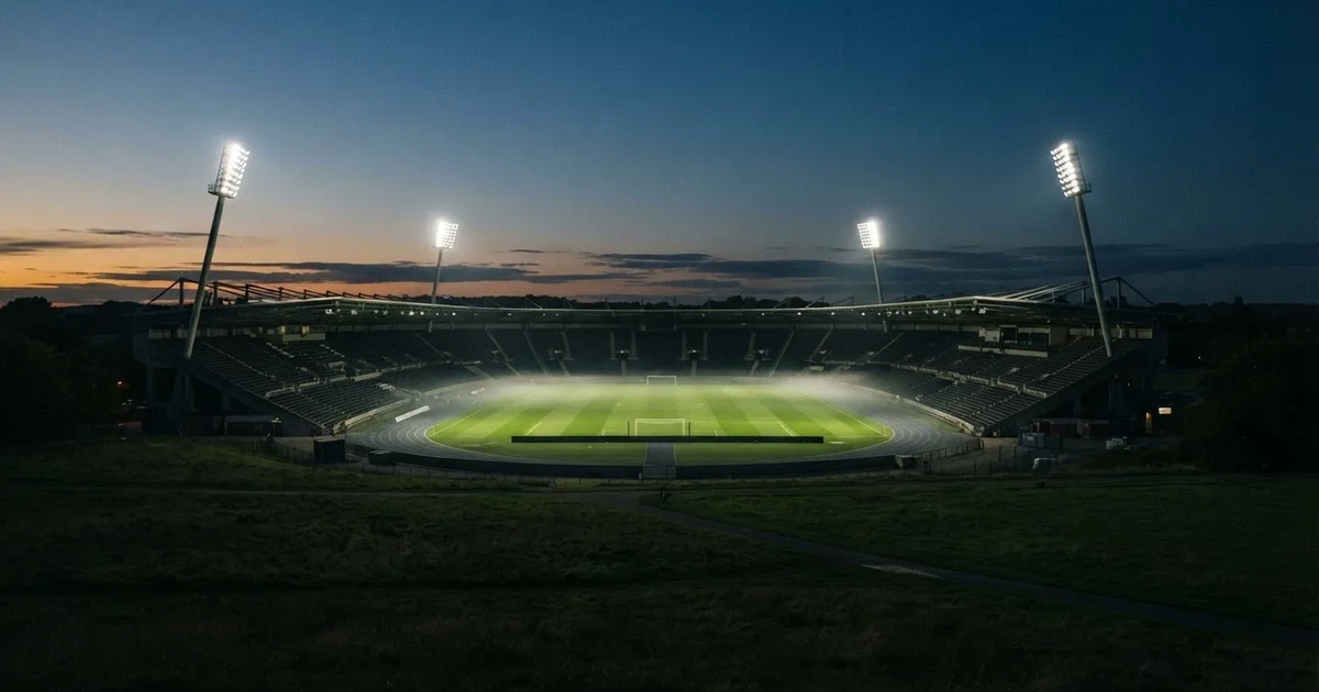 Ein Stadion unter Flutlicht aus der Ferne am Abend, hell erleuchtet, Nebel liegt über dem Rasen.
