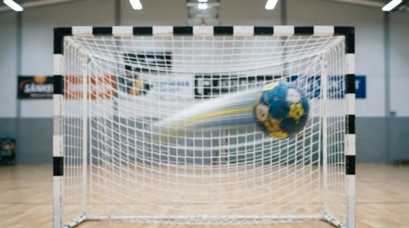 Ein Handball schwebt vor einem Tor mit Netz, die Szene zeigt Bewegungsunschärfe unter hellem Hallenlicht, keine Personen.