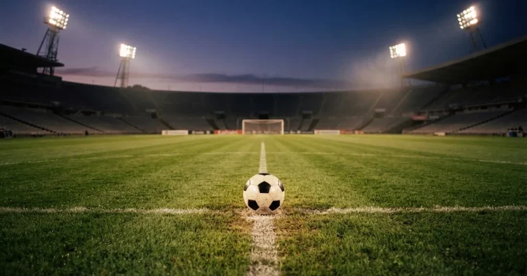 Ein Fußball liegt auf dem Anstoßpunkt eines Rasenplatzes in der Dämmerung, in der Ferne leuchten Flutlichter, die Ränge sind leer.