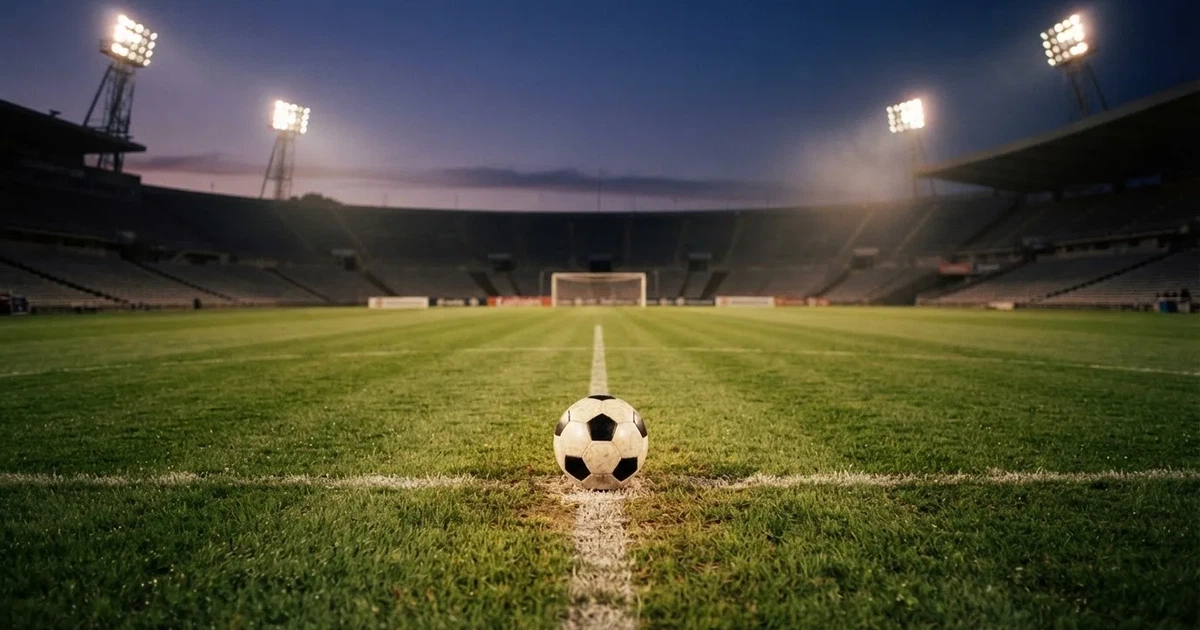 Ein Fußball liegt auf dem Anstoßpunkt eines Rasenplatzes in der Dämmerung, in der Ferne leuchten Flutlichter, die Ränge sind leer.